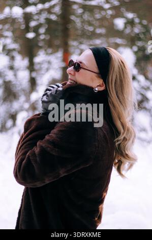 Femme aux longs cheveux blonds portant des lunettes de soleil et un manteau de fourrure brun sourit tout en se tenant dans une forêt enneigée Banque D'Images