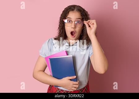 Élève surpris tenant des cahiers et des lunettes avec un fond rose dans une salle de classe Banque D'Images