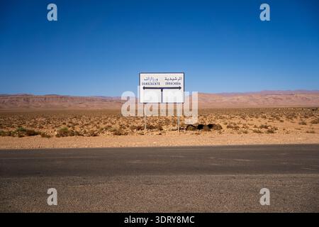 Une vue saisissante et minimaliste d’un panneau routier blanc bilingue (arabe et français) pointant vers Ouarzazate et Errachidia. Situé le long d'un a solitaire Banque D'Images