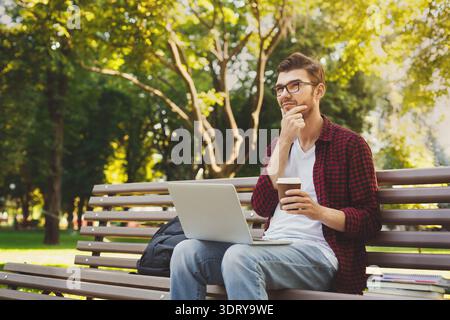 Homme assis sur le banc travaillant sur ordinateur portable dans le parc avec café à la main et expression réfléchie Banque D'Images