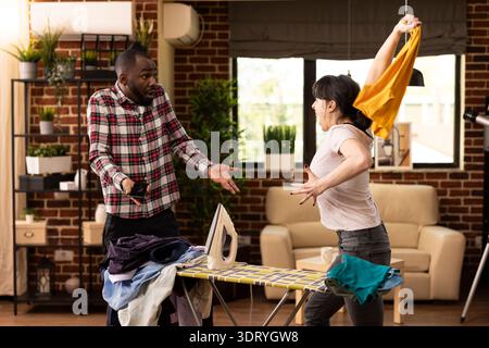 Femme blanche exprimant sa frustration pendant la journée de lessive, jetant une chemise vers un petit ami afro-américain. L'homme noir répond avec un geste confus pendant la dispute avec la petite amie dans un appartement moderne. Banque D'Images