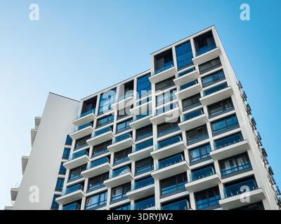 Bâtiment résidentiel blanc moderne contre le ciel bleu clair. Vue sous-jacente d'appartements contemporains de grande hauteur à façade géométrique avec balcons en verre. Banque D'Images