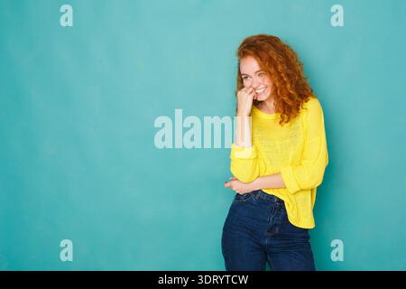 Femme souriante tout en se tenant debout contre un mur bleu avec une pose ludique et portant un pull jaune Banque D'Images