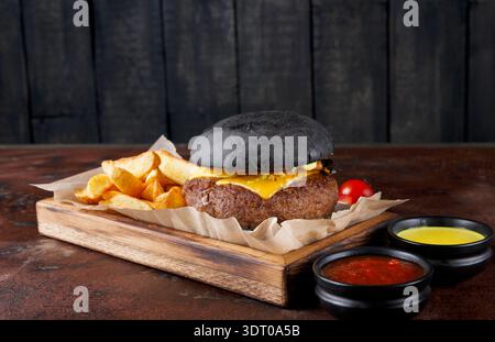 Burger Black Bun avec fromage et frites servi avec des trempettes sur une planche de bois dans un restaurant Banque D'Images