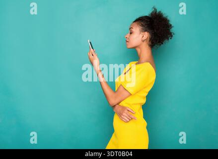 Jeune femme regarde son téléphone tout en se tenant debout contre un mur Turquoise dans une tenue lumineuse Banque D'Images