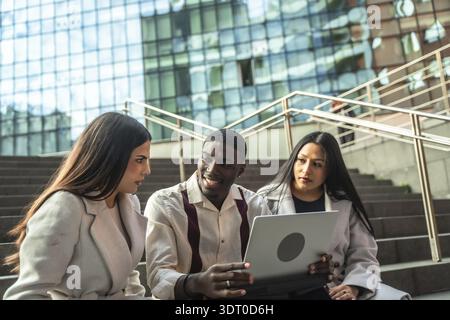 Divers professionnels des affaires ou étudiants assis dans les escaliers urbains, discutant des idées de projet et partageant des informations sur un ordinateur portable lors d'une ou informelle Banque D'Images