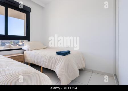 Chambre contemporaine lumineuse avec deux lits simples, literie neutre douce, grande fenêtre et carrelage propre dans un appartement moderne. Banque D'Images