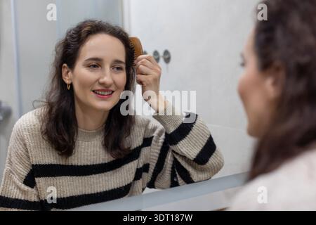 Femme souriante se brossant les cheveux devant un miroir, routine matinale décontractée dans une salle de bain lumineuse. Banque D'Images