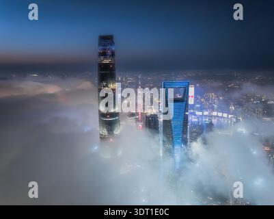 Vue aérienne de gratte-ciel perçant à travers l'épaisse couverture de brouillard, leurs lumières se reflétant contre l'air brumeux, Shanghai, Chine. Banque D'Images