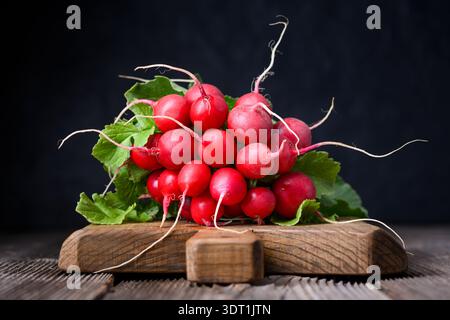 Radis rouges frais avec des dessus feuillus sur planche de bois dans un éclairage discret. Légumes naturels pour le régime détox et le style alimentaire rustique Banque D'Images