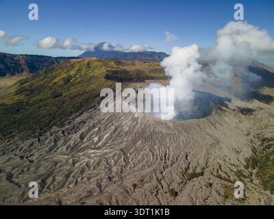 Vue aérienne du cratère dur du mont bromo exhalant des panaches de fumée blanche sur fond de terrain accidenté et de pics lointains, le mont bromo, East Jav Banque D'Images