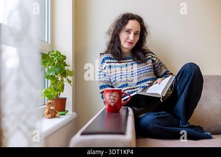 Femme d'âge moyen se relaxant avec un livre et une tasse. Portrait confortable de style de vie d'une femme brune d'âge moyen assise sur un canapé près de la fenêtre, lisant un livre Banque D'Images