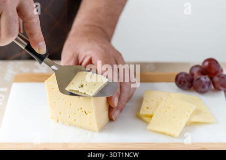 Un homme dans un tablier sombre coupant un morceau de fromage avec une trancheuse spéciale une planche de marbre dans la cuisine. Fond clair avec espace de copie Banque D'Images