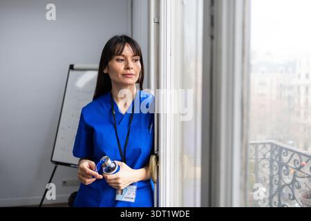 Un médecin en gommages bleus se tient près d'une fenêtre, regardant soigneusement à l'extérieur. Elle tient une longe et une bouteille d'eau, représentant l'engagement et la préparation i. Banque D'Images