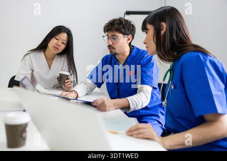 Une équipe médicale professionnelle composée de deux médecins en gommages bleus et une infirmière en vêtements blancs ayant une discussion autour d'une table dans un clinique Banque D'Images