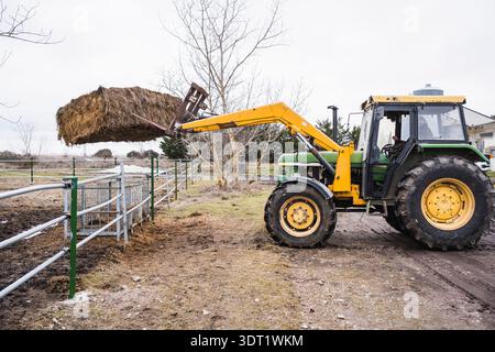 Un tracteur équipé d'un chargeur frontal lève de grosses balles de foin, placées contre un paysage agricole rural. La scène illustre le rôle des machines dans l'agriculture Banque D'Images