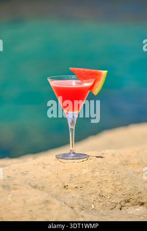 Cocktail de pastèque dans un verre à martini assis sur un rocher avec la mer en arrière-plan. Banque D'Images
