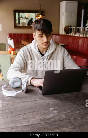 Un jeune homme concentré sur son ordinateur portable à une table de café moderne, avec des écouteurs élégants à proximité, incarne le mélange de technologie et de style décontracté Banque D'Images