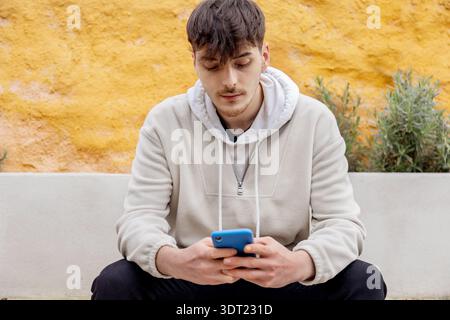 Un jeune homme vêtu de façon décontractée d'un sweat à capuche regarde son smartphone assis contre un mur jaune éclatant à l'extérieur, mettant en valeur la technologie moderne Banque D'Images