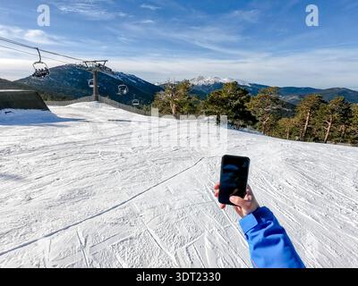 Un skieur tient un smartphone sur le fond enneigé de Puerto de Navacerrada, Madrid, avec des remontées mécaniques qui montent les montagnes enneigées pittoresques Banque D'Images