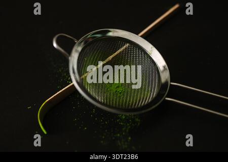 Une image en gros plan de poudre de matcha vert éclatant dans un tamis métallique reposant sur une surface noire. La scène capture l'essence du thé traditionnel pré Banque D'Images