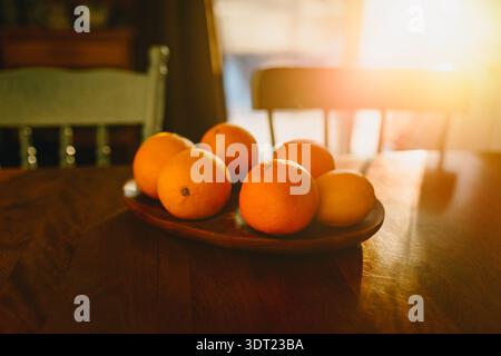 Un bol en bois rempli d'oranges vibrantes se trouve sur une table rustique, illuminée par la lueur chaude du soleil du matin, créant un kitche confortable et accueillant Banque D'Images