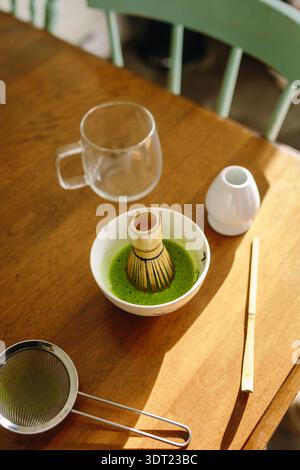 Une installation sereine pour préparer du thé matcha, avec un fouet en bambou immergé dans du matcha vert vif, une tasse en verre, une passoire et une cuillère sur un t en bois Banque D'Images
