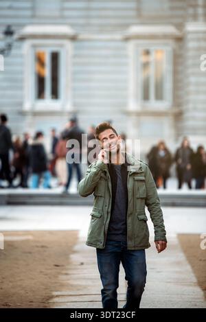 Un jeune homme en veste verte marche en toute confiance dans une rue animée de Madrid, en Espagne, discutant au téléphone avec une expression joyeuse, entouré Banque D'Images