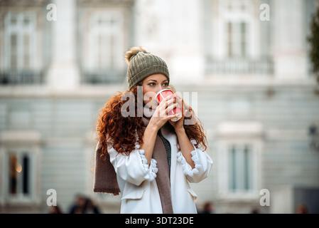 Une femme dans un manteau confortable et chapeau de laine savoure une boisson chaude d'une tasse rouge, debout à l'extérieur contre une façade de bâtiment classique à Madrid, Espagne sur un ch Banque D'Images