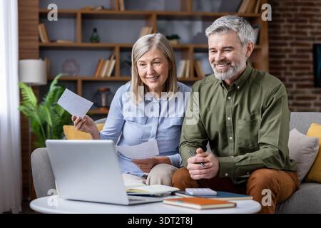 Couple senior assis sur un canapé dans leur salon, souriant et travaillant avec un ordinateur portable, gérant les finances de la maison, examinant les factures et discutant de la planification budgétaire mensuelle Banque D'Images