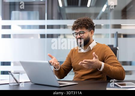 Jeune homme d'affaires avec des cheveux bouclés et des lunettes parlant lors d'une vidéoconférence sur ordinateur portable, partageant des idées et communiquant efficacement dans un environnement moderne Banque D'Images