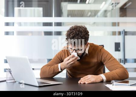 Homme avec des cheveux bouclés et des lunettes soufflant son nez dans un mouchoir tout en travaillant sur un ordinateur portable à son bureau dans un bureau moderne, représentant la santé au travail an Banque D'Images