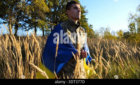 Homme militaire en uniforme se tient avec le drapeau de l'Ukraine sur les épaules à la campagne. Portrait de soldat ukrainien masculin avec la bannière nationale en l'honneur de Banque D'Images