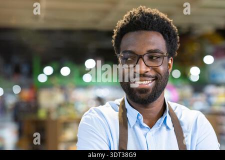 Homme joyeux portant des lunettes et un tablier, souriant avec confiance et regardant loin tout en se tenant debout dans son petit magasin d'entreprise, représentant le succès et l'esprit d'entreprise Banque D'Images