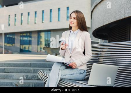 Femme d'affaires s'assoit sur un banc en utilisant un smartphone et en regardant des documents dans la zone urbaine Banque D'Images