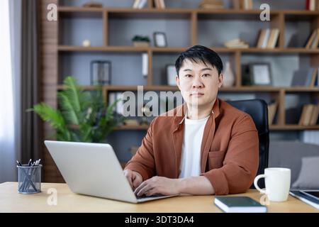 Homme asiatique assis à un bureau, regardant la caméra tout en tapant sur un ordinateur portable, entouré d'éléments de bureau, symbolisant la productivité, la concentration et le travail à distance dans un environnement confortable Banque D'Images