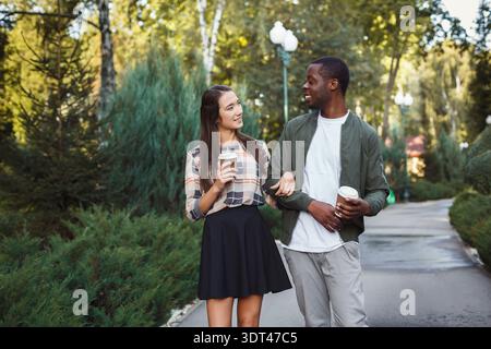Couple aime une promenade dans un parc tout en tenant des tasses de café et en souriant l'un à l'autre Banque D'Images