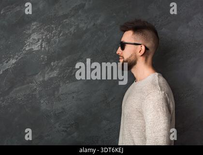 Homme portant des lunettes de soleil pose contre un mur texturé dans un cadre élégant avec des couleurs calmes Banque D'Images