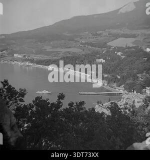 Une superbe photographie d'archives en noir et blanc des années 1960 offre une vue panoramique sur la plage bondée et le littoral de Gurzuf, en Crimée. Prise depuis un point de vue élevé, la scène présente un rivage animé rempli de vacanciers, une longue jetée et un bateau de plaisance blanc naviguant dans les eaux calmes de la mer Noire. Des collines luxuriantes avec des sanatoriums soviétiques emblématiques et une végétation méditerranéenne dense s'élèvent en arrière-plan. Cette image historique capture parfaitement l'âge d'or du tourisme soviétique et l'atmosphère sereine de la station balnéaire du milieu du XXe siècle. Banque D'Images