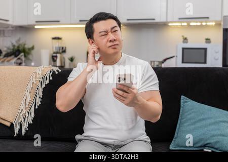 Asiatique inspiré homme à la maison études en ligne sur smartphone porte des écouteurs entend la lecture et trouve la réponse. Le gars chinois sur le canapé montre OK signe fixe l'objectif pour la semaine sauve la tâche à l'application sourires commence le plan Banque D'Images