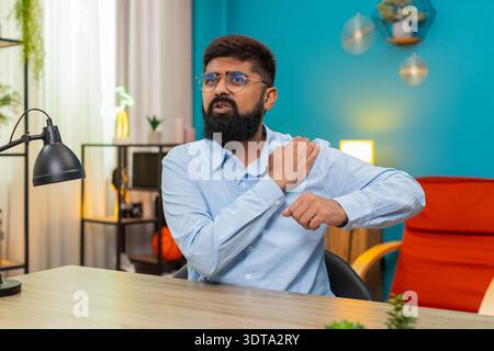Homme indien à la maison griffes épaule griffant de douleur aiguë sévère possible blessure inconfort musculaire tendu. Arabian Guy au bureau grimaces à la recherche de massage et de repos espoirs pour la douleur de récupération facile bientôt. Banque D'Images