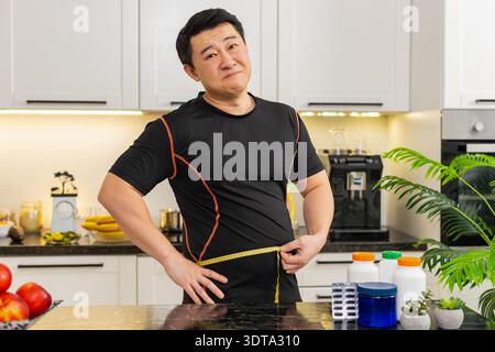 L'homme asiatique en surpoids dans la cuisine à la maison mesure la taille avec du ruban adhésif et semble frustré par le gros ventre après le gain de poids. Les capsules de régime sur la table suggèrent la recherche pour le changement de routine de santé de soutien amincissant. Banque D'Images