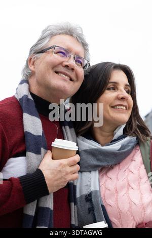 Couple d'âge moyen debout dehors sous le ciel nuageux, tenant des tasses à emporter et portant des foulards Banque D'Images