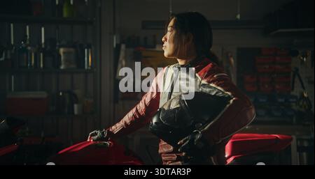 Déterminé jeune femme asiatique en costume d'équitation en cuir se tient à côté de Red Sport Motorcycle à l'intérieur élégant garage industriel. L'esprit intrépide des cavalières modernes embrassant la vitesse et l'indépendance. Banque D'Images