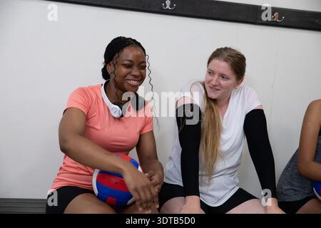 Diverses coéquipières féminines assises sur un banc de gymnastique tenant des balles de volley-ball rouges bleues et des écouteurs Banque D'Images