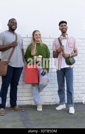 Divers amis appuyés contre le mur de briques peintes en blanc sur l'asphalte, tenant le café, les sacs, le téléphone Banque D'Images