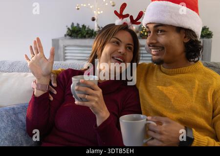 Couple diversifié assis sur le canapé à la maison, tenant des tasses, portant chapeau de Père Noël et serre-tête en bois Banque D'Images