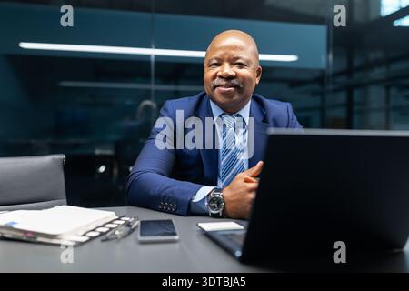 Homme d'affaires noir souriant tout en étant assis au bureau avec ordinateur portable dans le bureau moderne Banque D'Images