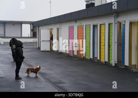 Cabanes de plage colorées à Littlehampton avec personne promenant un petit chien sur le parvis humide, portes vibrantes et ciel couvert, côte ouest du Sussex Angleterre. Banque D'Images