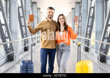 Couple heureux tenant des passeports et souriant au pont de l'aéroport, debout avec des valises Banque D'Images